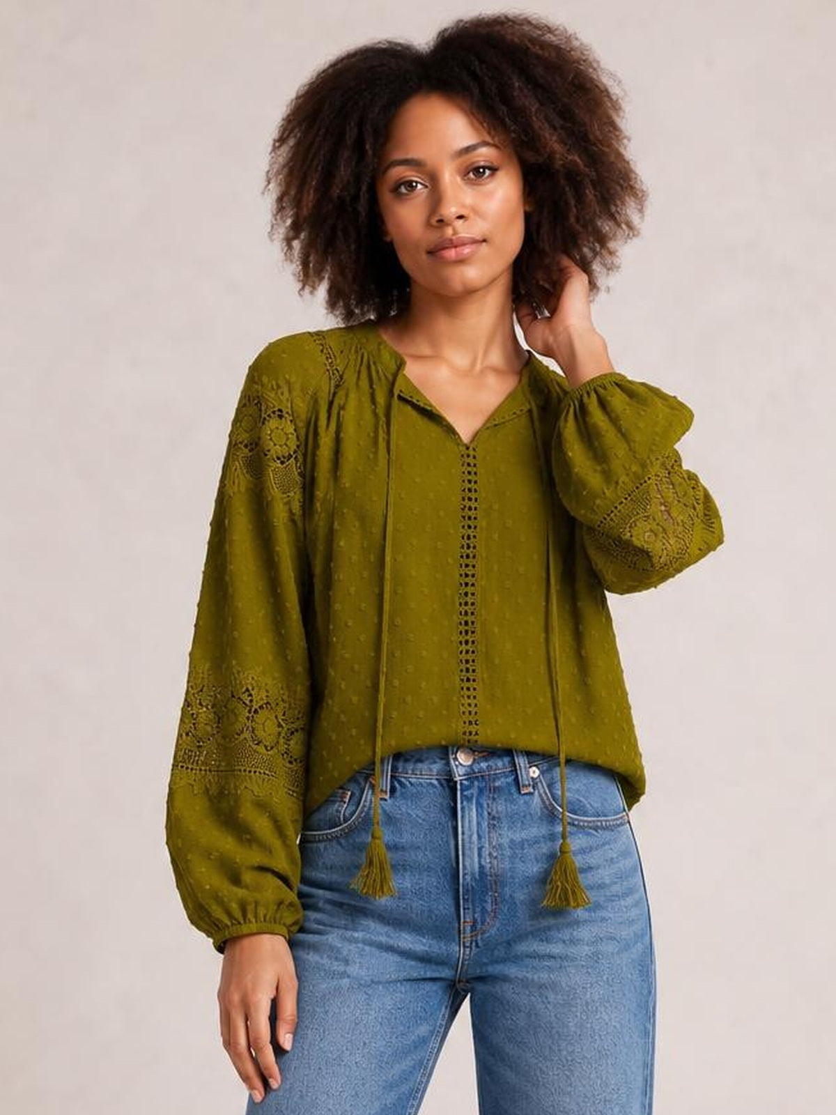 Model wears an olive green crochet-trim blouse with a tie neckline, tassel ends, long voluminous sleeves, and a relaxed silhouette while touching her hair.