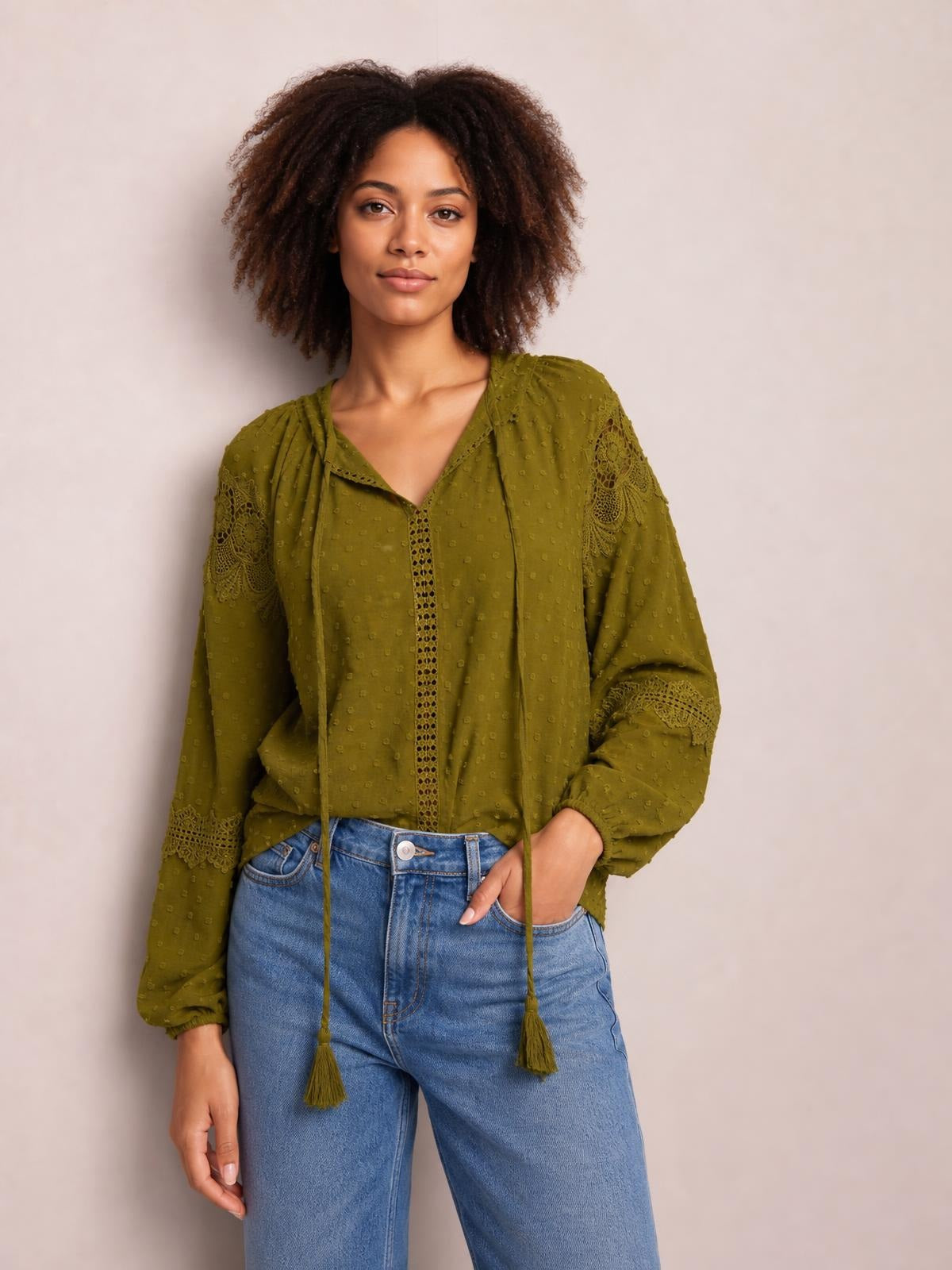 Model poses in an olive-green crochet tassel tie-neck blouse with long sleeves, a relaxed silhouette, dotted texture, and lace-like sleeve inserts.