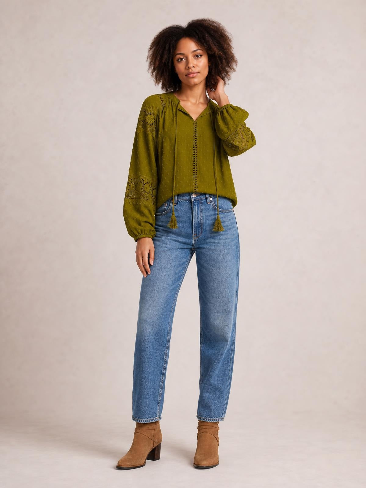 A model poses in an olive green crochet blouse with a tie neck, tassel ends, long sleeves, lace panel details, and a relaxed silhouette.