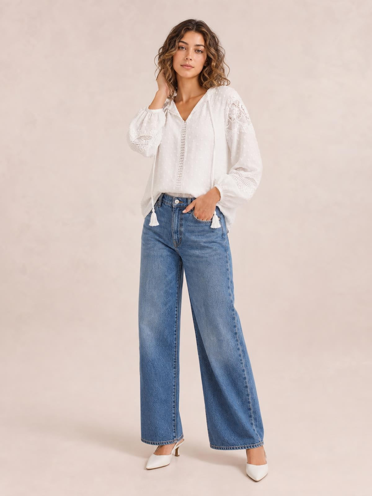 A model poses in a white crochet tassel tie-neck blouse with long sleeves, a relaxed silhouette, V-neckline, and semi-sheer lace detailing.