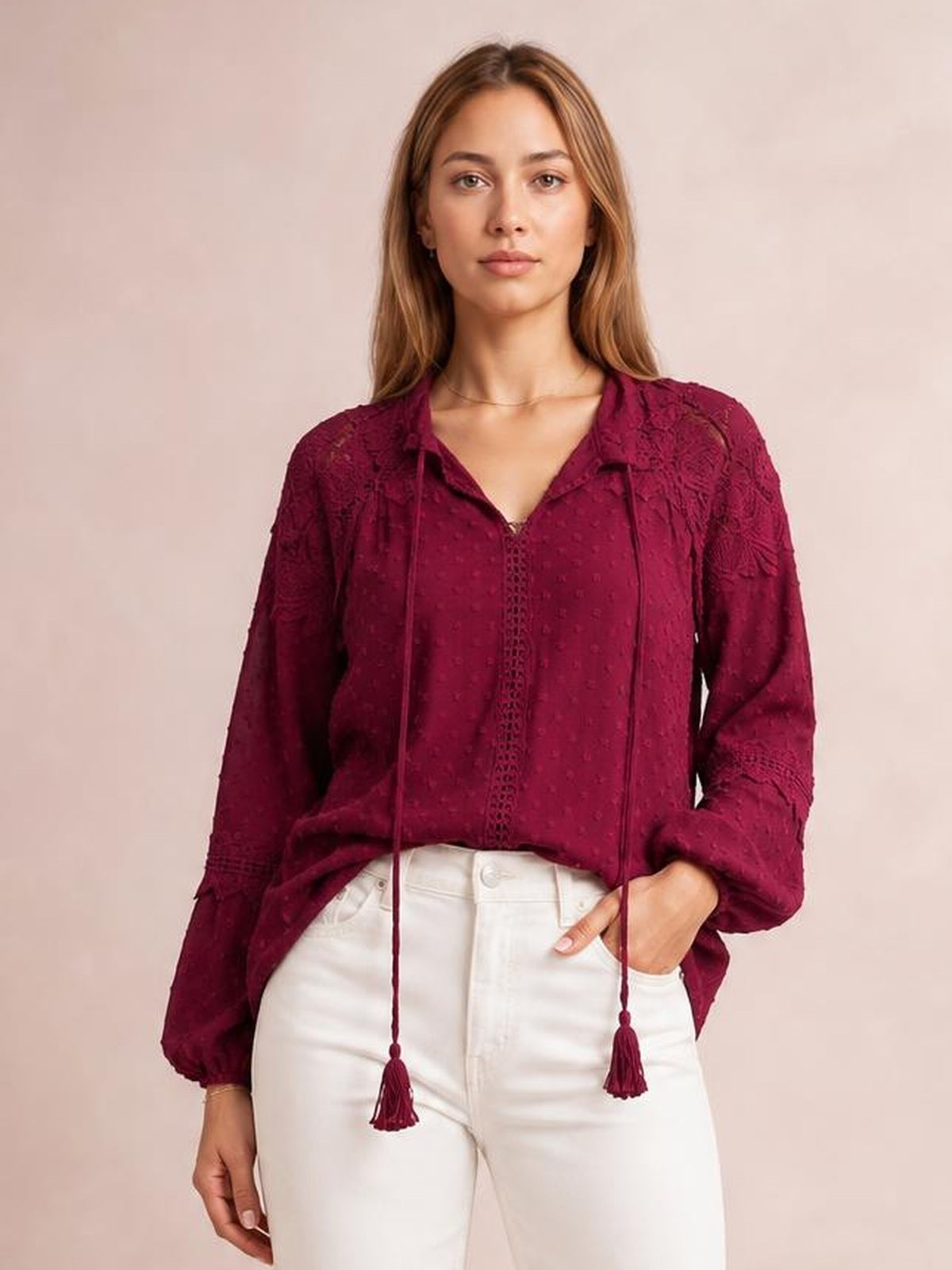 A model wears a burgundy crochet blouse with a tie neck, long sleeves, tassel ends, semi-sheer texture, and a relaxed silhouette.