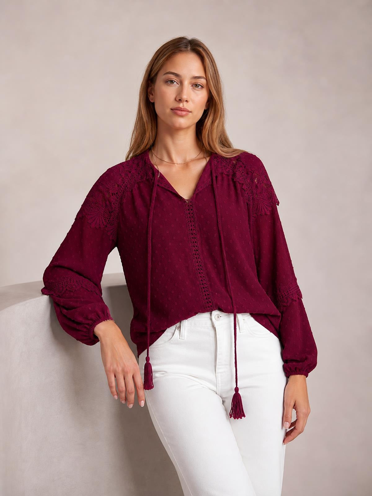 A model leans against a wall wearing a burgundy crochet tassel tie-neck blouse with long sleeves, lace panels, elastic cuffs, and a relaxed silhouette.
