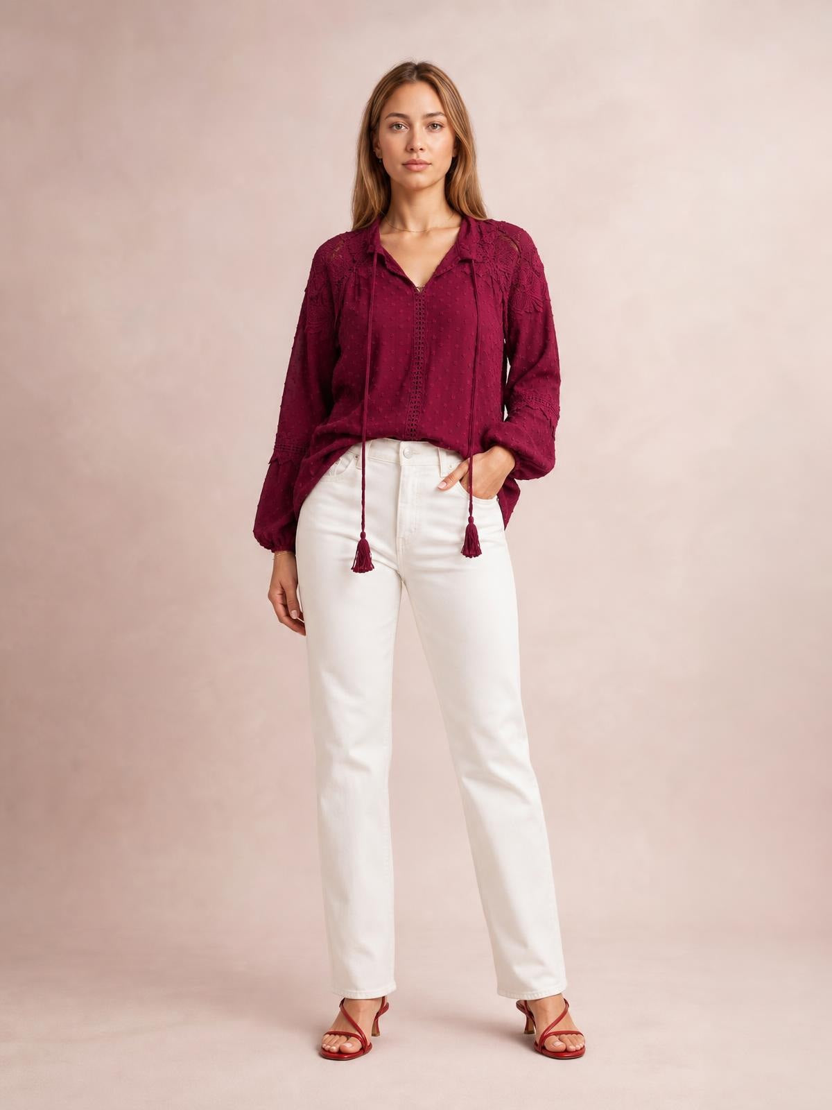 A model wears a burgundy crochet blouse with a tie neckline, long tassel ties, textured sleeves, and a relaxed silhouette while standing with one hand in her pocket.