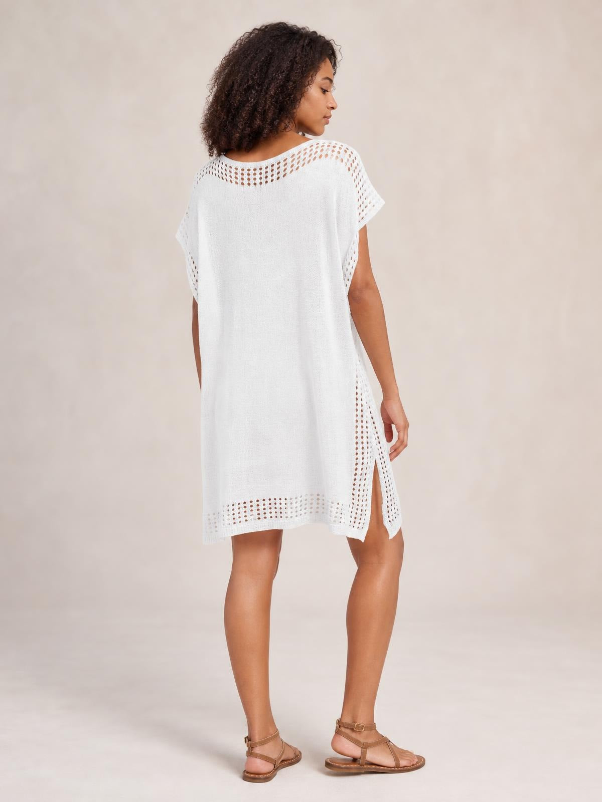 Model turned slightly away wears a white beach cover up dress with a loose silhouette, crochet-trim cutouts, short sleeves, and a side slit.