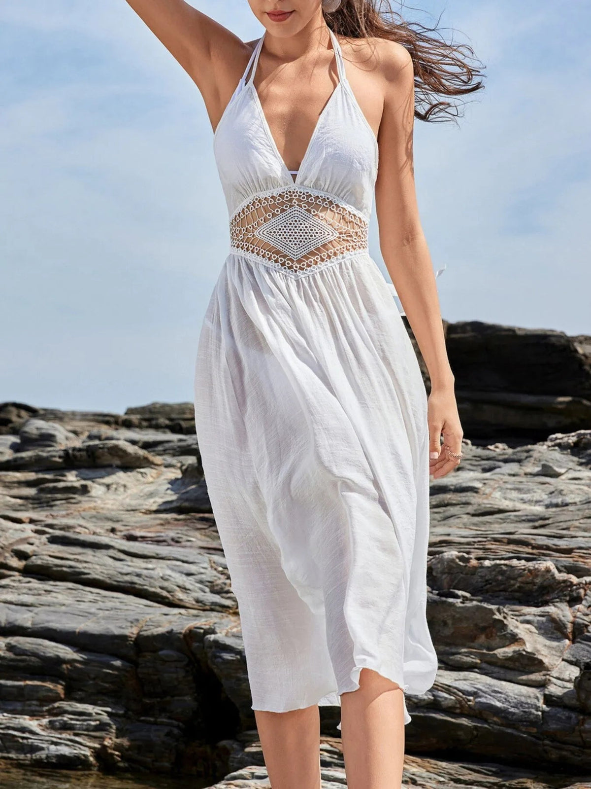 Model raises one arm wearing a white crochet waist halter sleeveless cover-up dress with tied straps, cutout waist, gathered bodice, and flowing silhouette.