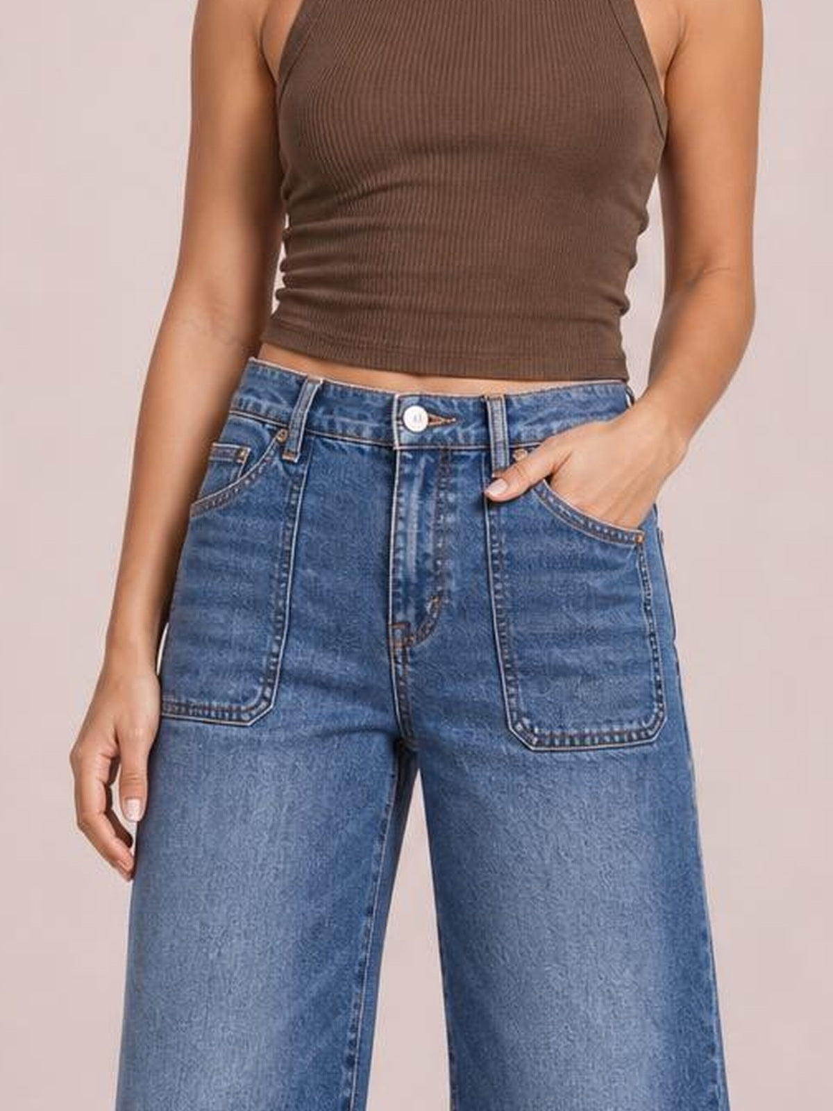 A model wears blue cropped high-rise wide-leg jeans with contrast stitching, large front patch pockets, belt loops, and one hand tucked into a pocket.