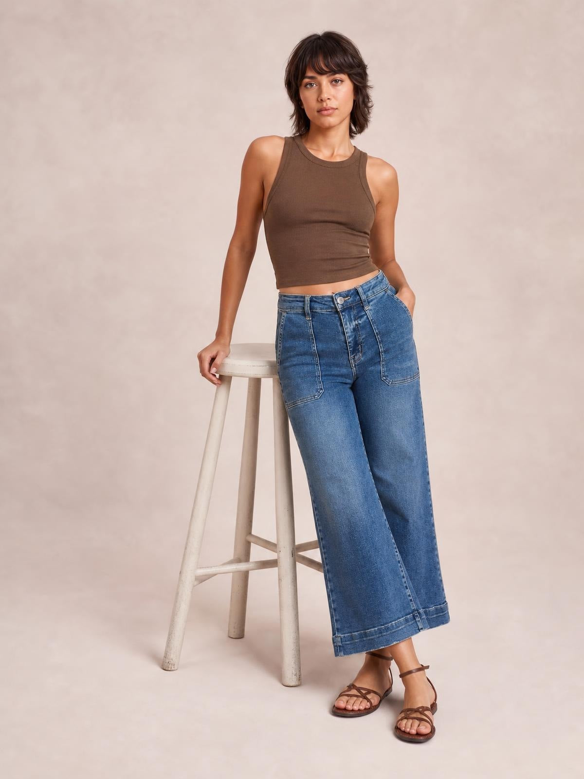 Model leans on a stool wearing blue cropped high-rise wide-leg jeans with front and side pockets, belt loops, a button fly, and wide cuffed hems.