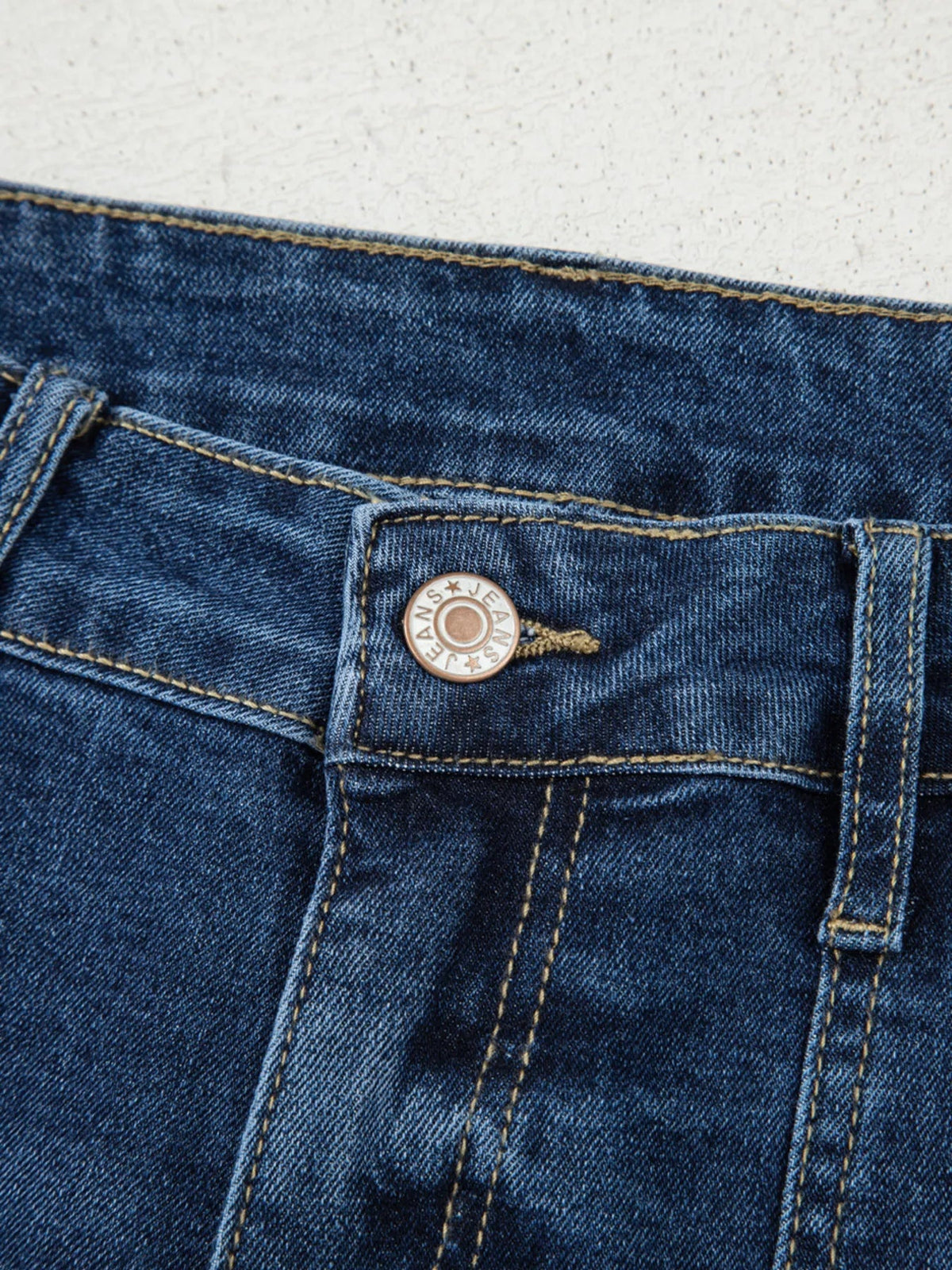Close-up of dark blue high-rise jeans showing the waistband, pocket, metal button, belt loop, and contrasting golden stitching.