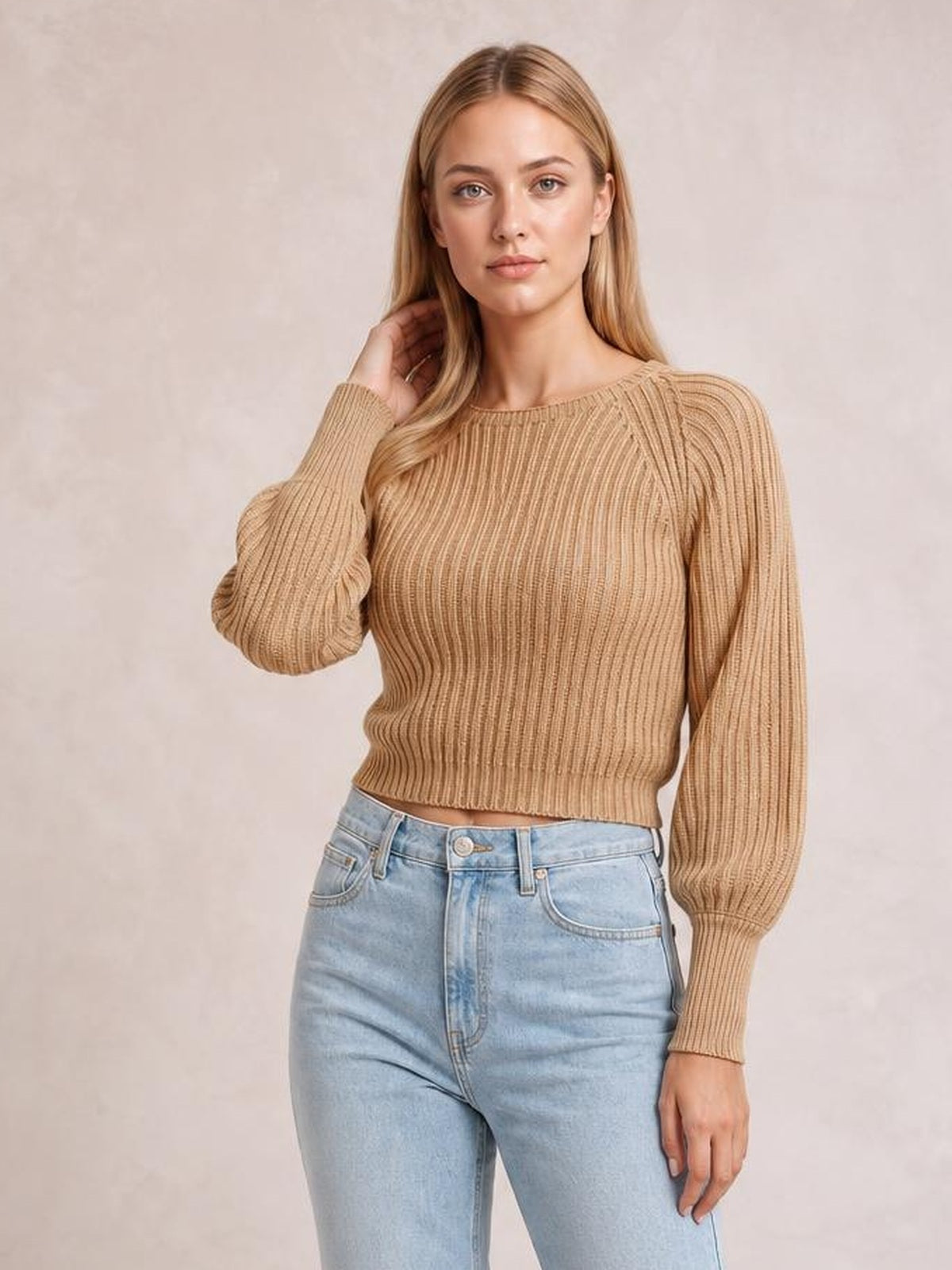 A model poses in a camel cropped ribbed pullover sweater with a round neck, long raglan sleeves, fitted silhouette, and one hand touching her hair.