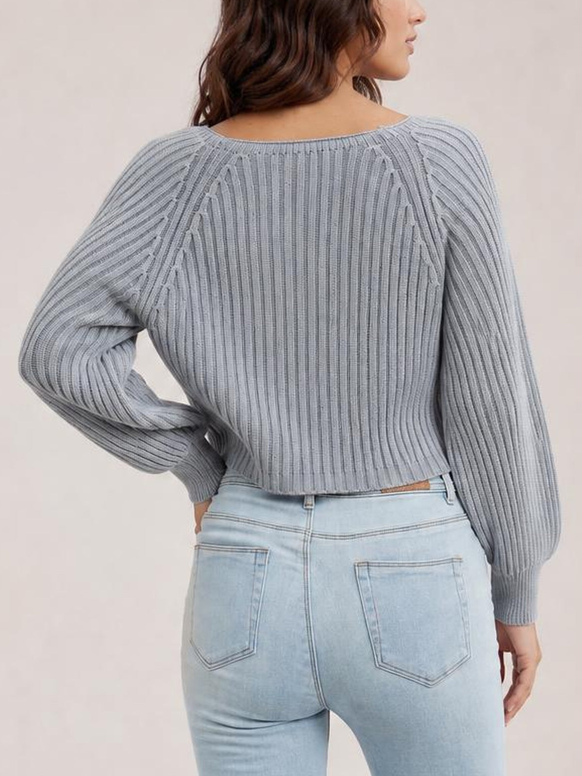 Model shown from behind wearing a solid gray cropped round-neck ribbed pullover sweater with long raglan sleeves, one hand resting on her hip.
