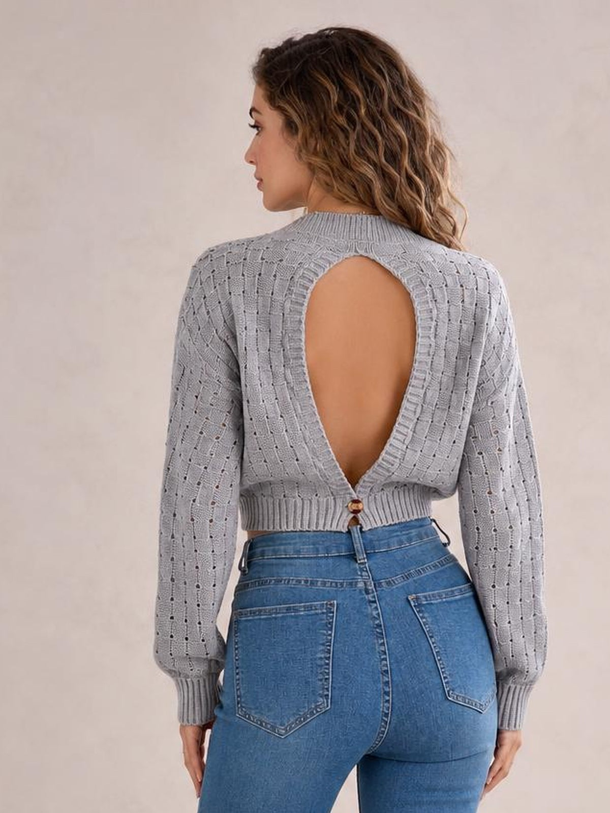 A model faces away to show a gray round-neck long-sleeve sweater with openwork knit texture, ribbed cuffs and hem, and a large buttoned cutout back.