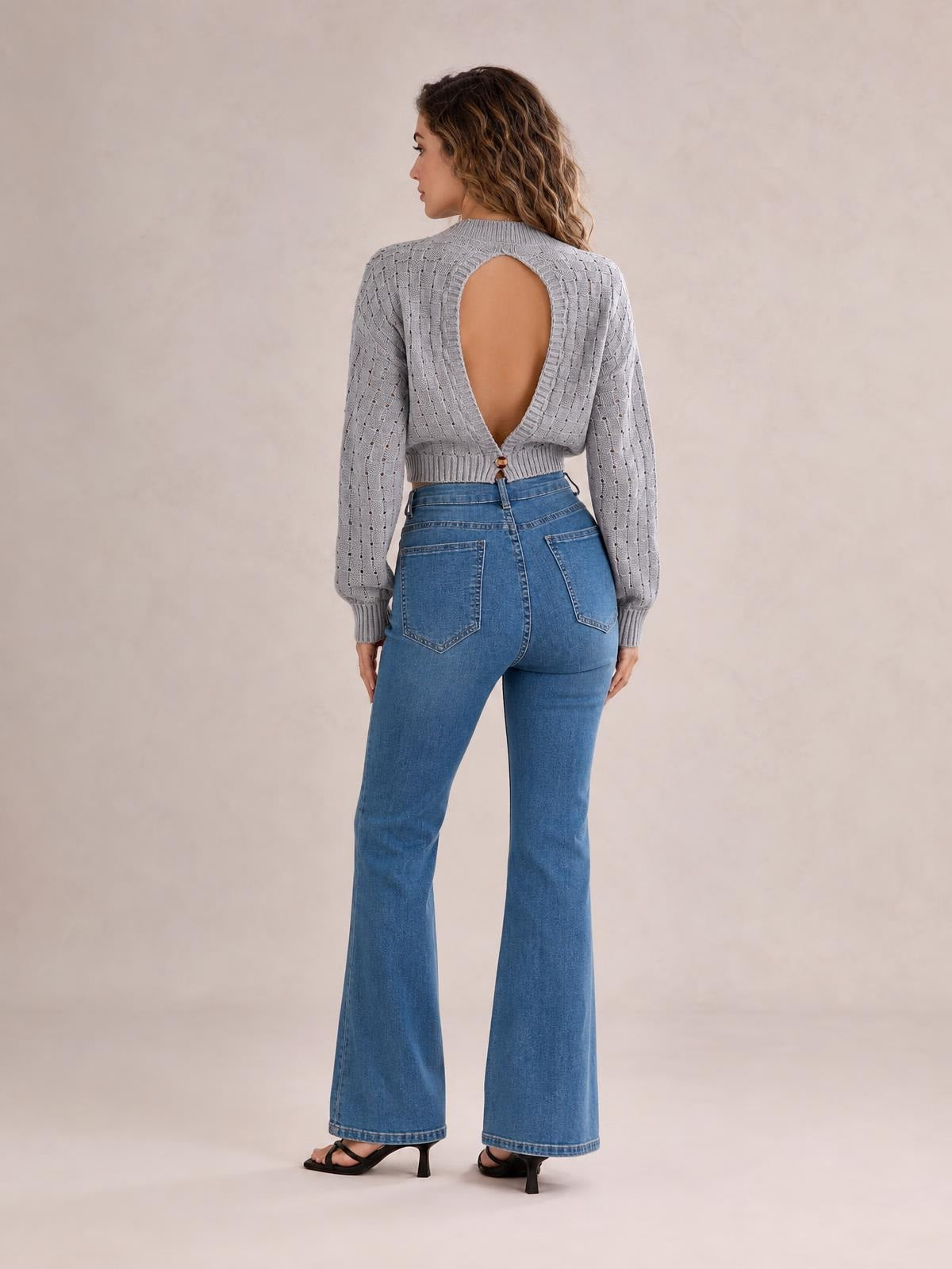 Model shown from behind in a light gray openwork sweater with long sleeves, a round neck, cropped fitted silhouette, and large cutout back with button closure.