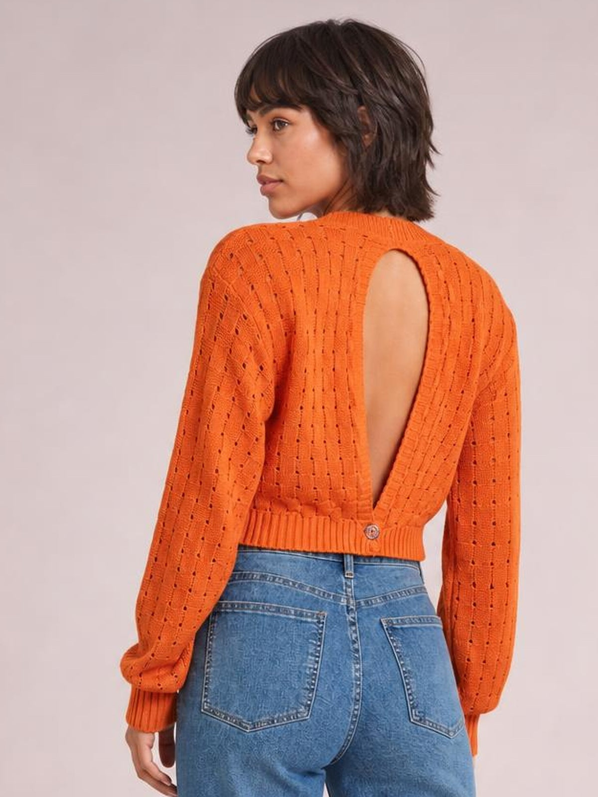 Model turns to show an orange openwork sweater with a round neck, long sleeves, ribbed hem and a large back cutout with button detail.