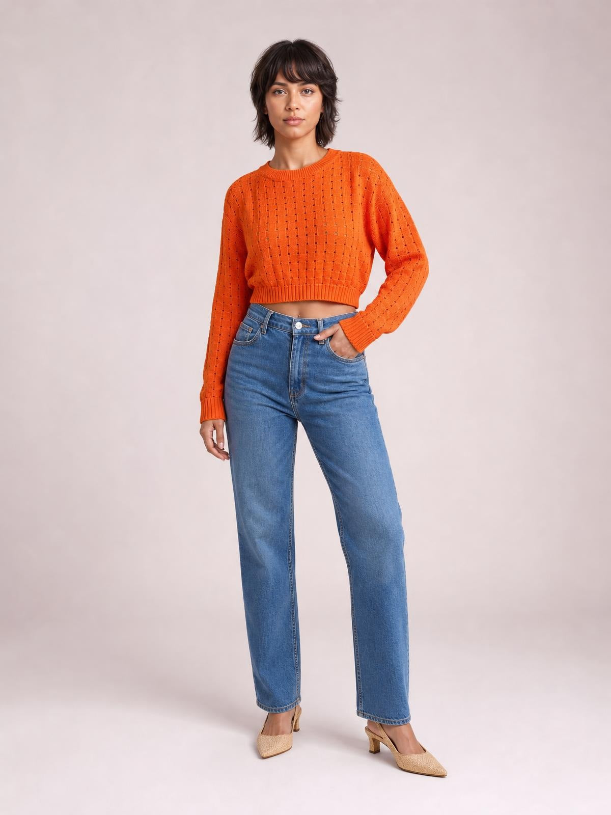 Model wears a bright orange openwork sweater with a round neck, long sleeves, ribbed cuffs and hem, cropped relaxed silhouette, and one hand in her jeans pocket.