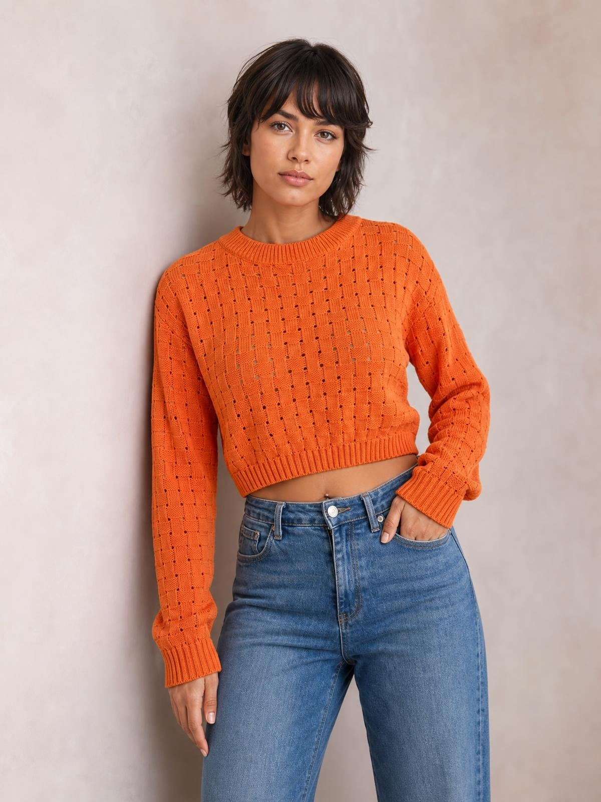 A model poses in an orange openwork sweater with a ribbed round neck, long sleeves, cropped hem, and ribbed cuffs and waistband.