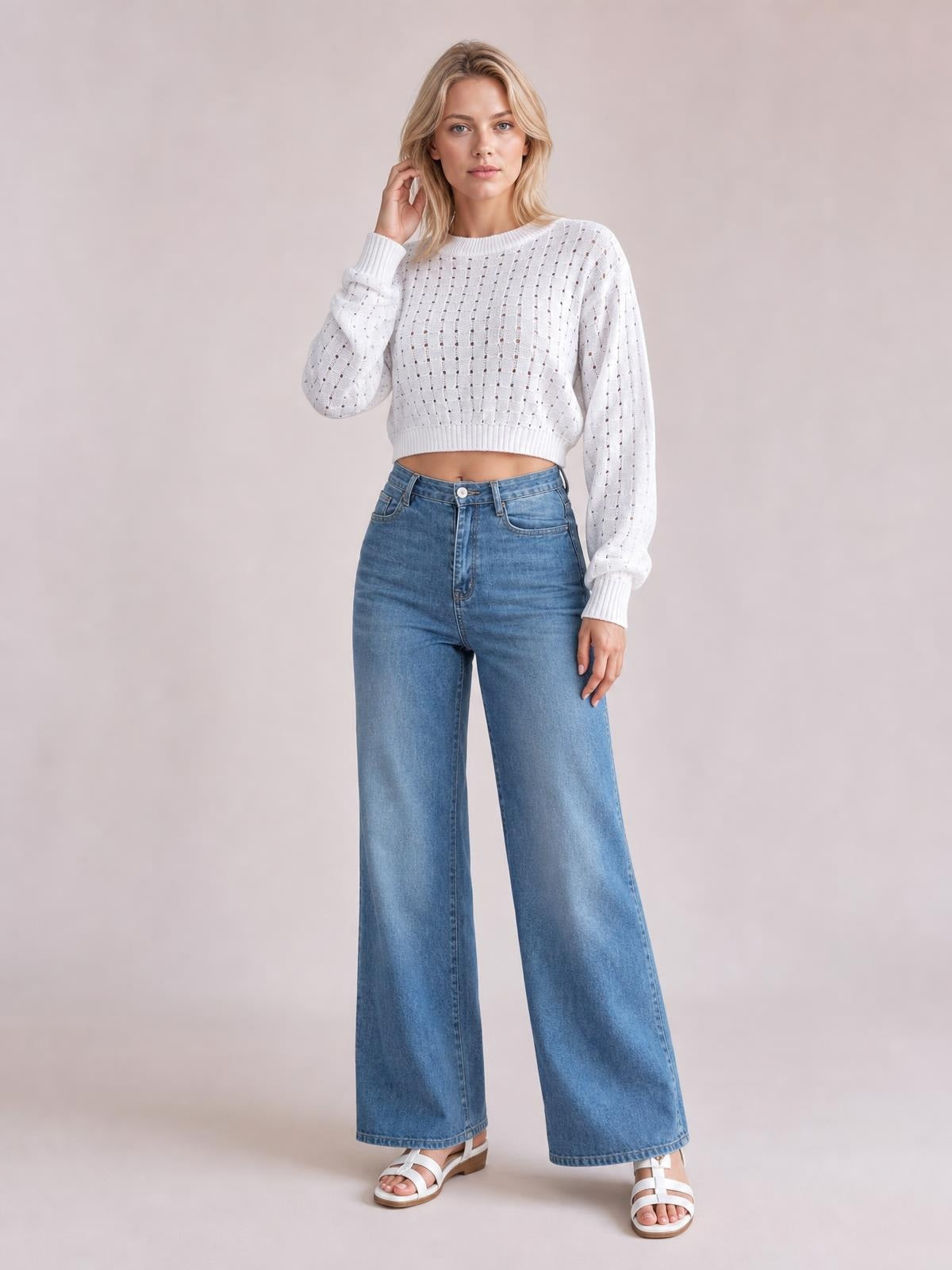 Model wears a white openwork sweater with a round neck, long sleeves, ribbed trim, and a cropped silhouette, posing with one hand near her hair.
