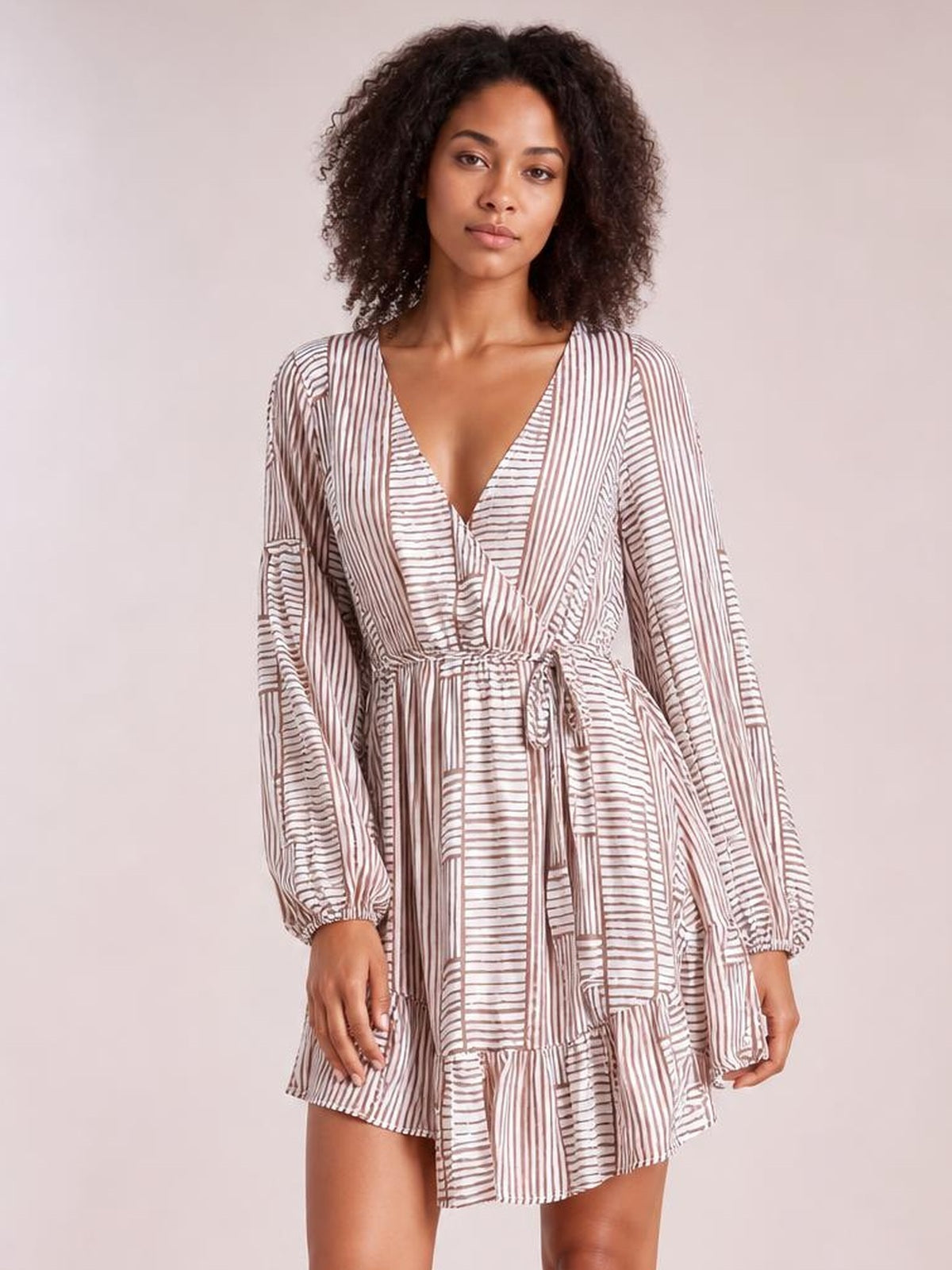 Model poses in a cream and brown striped surplice dress with long sleeves, a tied waist, relaxed silhouette, and ruffled tiered mini hem.
