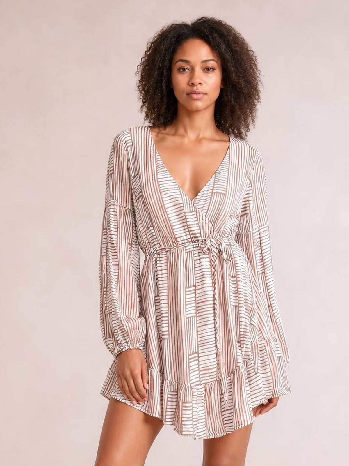 A model wears a cream and brown striped surplice long sleeve dress with a tied waist, relaxed short silhouette, and ruffled hem.