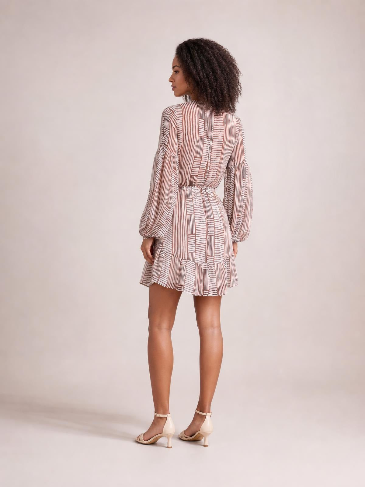 Model stands turned away in a white and brown striped dress with long puff sleeves, a tied waist, relaxed mini silhouette, and ruffled hem.