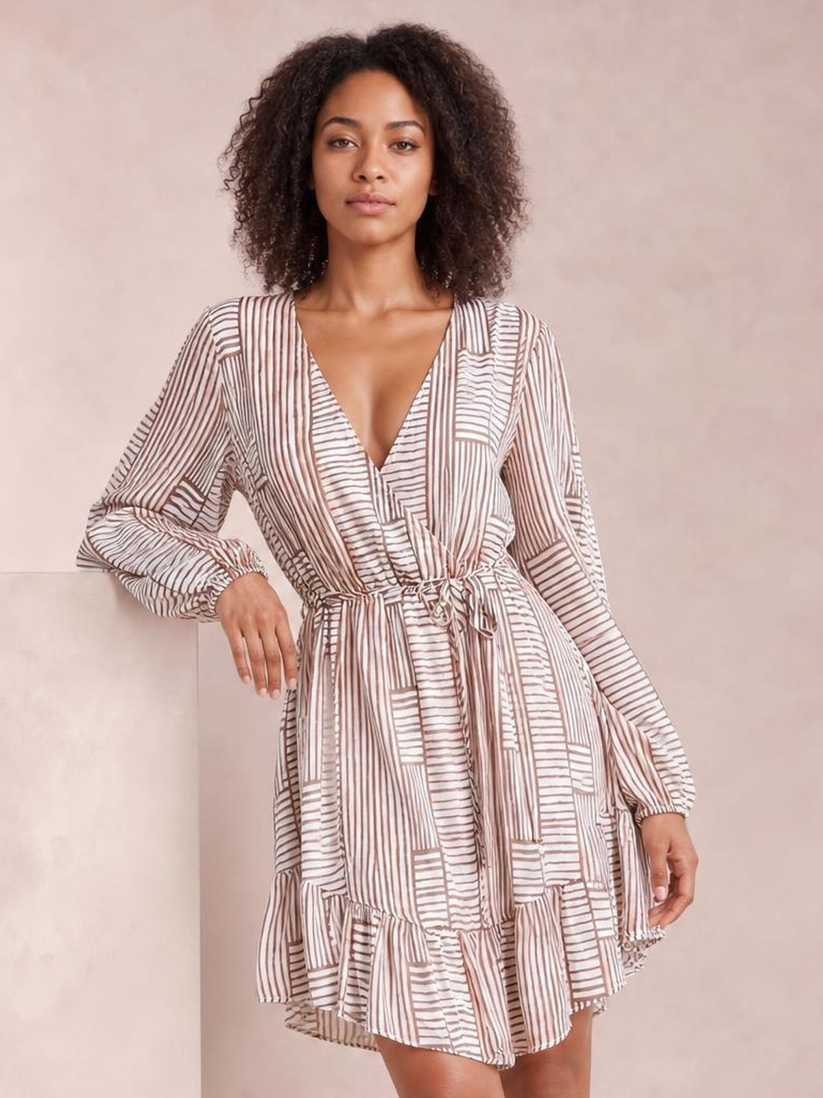 A model poses in a cream and brown striped surplice dress with a V-neck, long sleeves, tied waist, flared silhouette, and ruffled tiered hem.