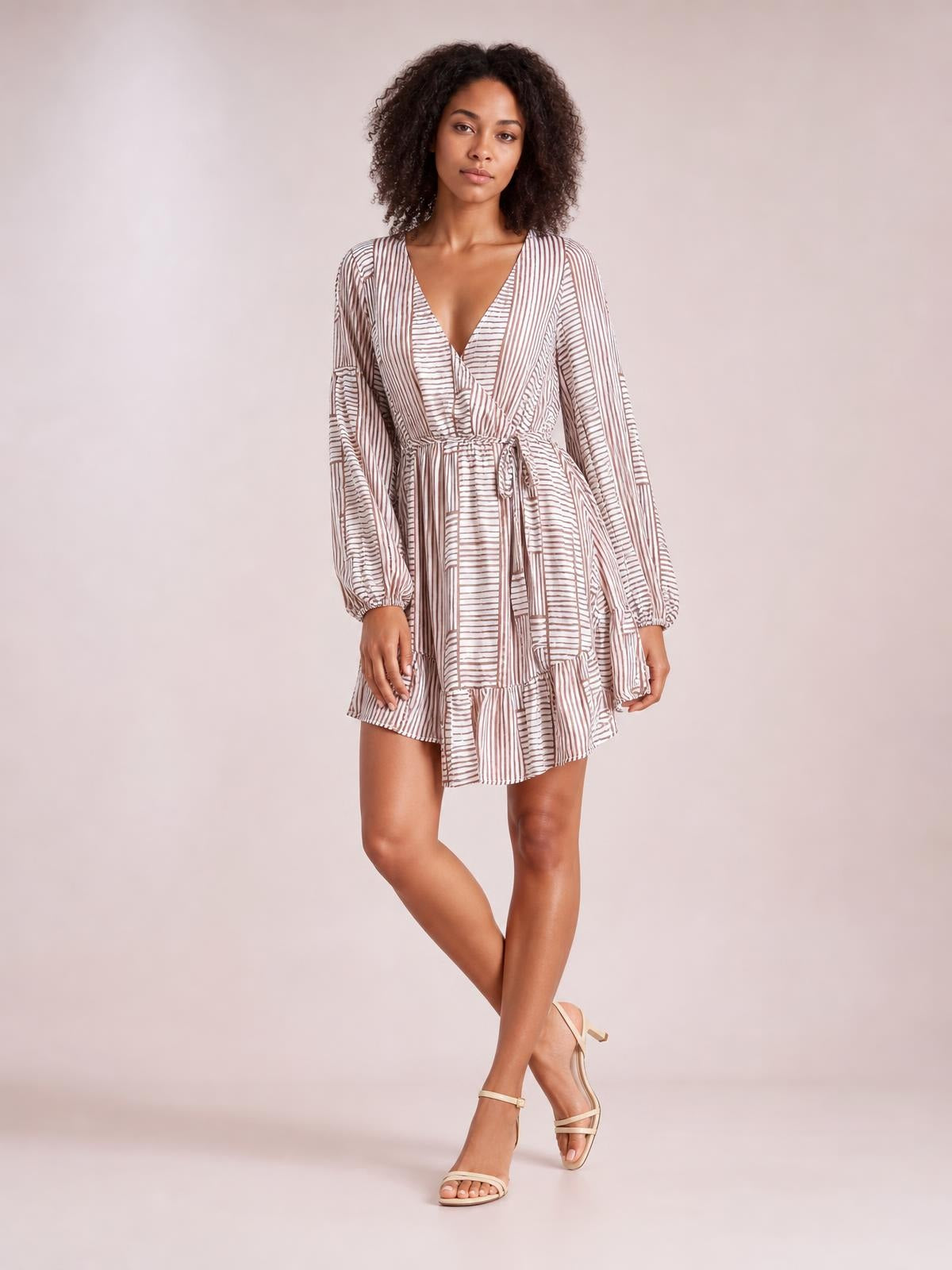 Model poses with crossed legs in a striped surplice mini dress featuring a V-neckline, long sleeves, tied waist, and tiered ruffled A-line silhouette.