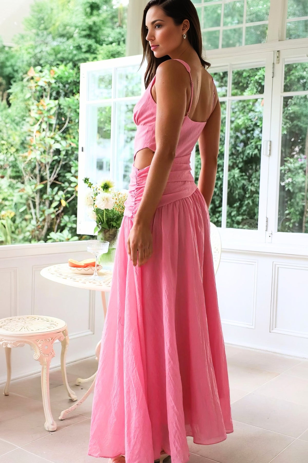 Model turned to show the back of a pink sleeveless V-neck dress with adjustable straps, ruched waist, side cutout, and flowing floor-length skirt.
