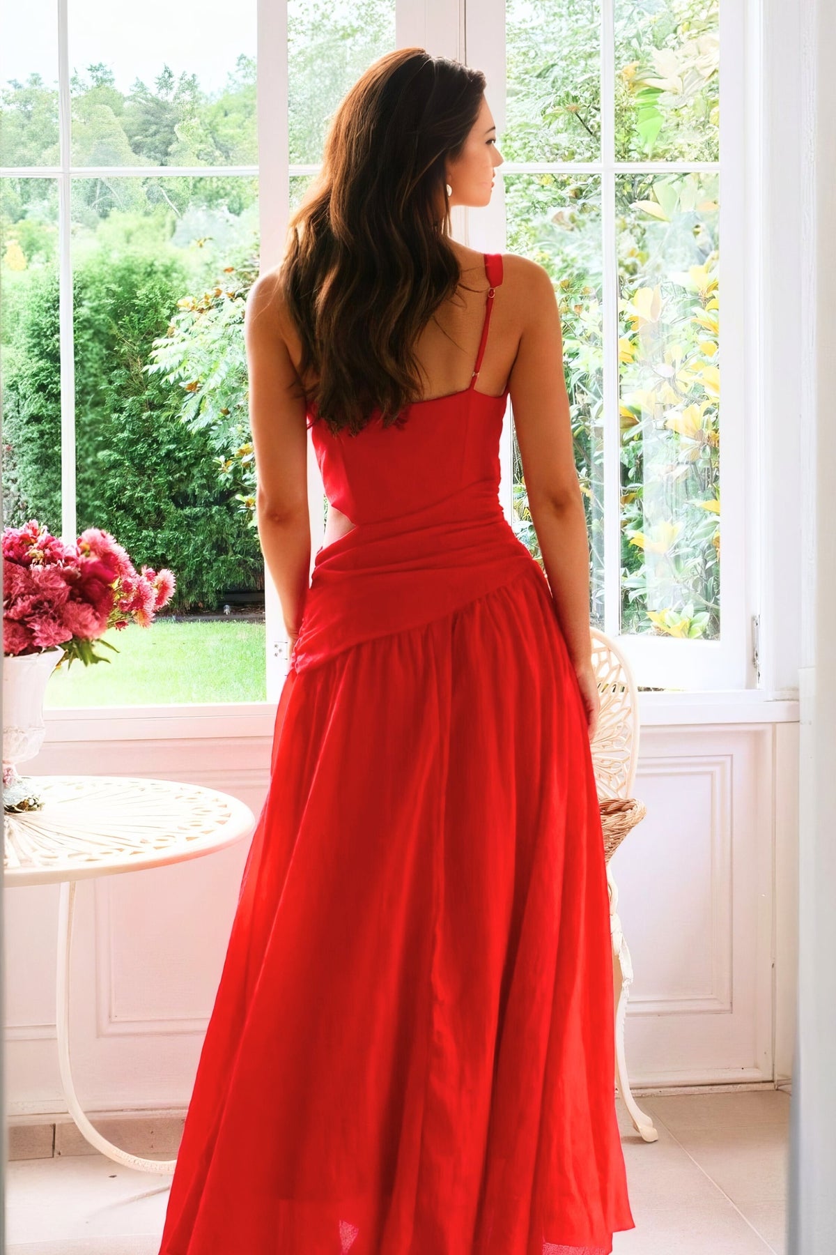 Model shown from behind in a red sleeveless dress with thin straps, a cutout back, ruched waist, and a flowing floor-length skirt.