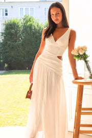 A model leans in an ivory sleeveless V-neck dress with ruched waist detailing, a fitted bodice, cutout side, and flowing floor-skimming skirt.