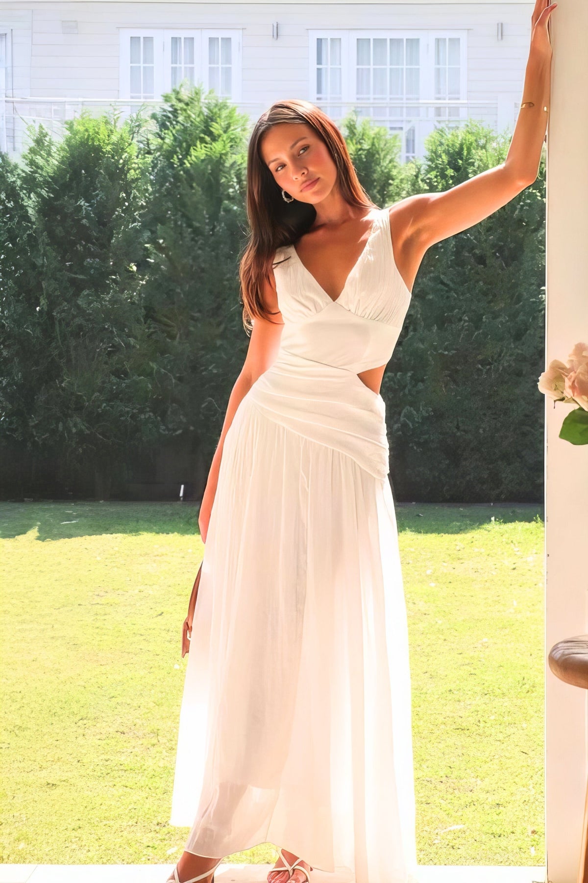 A model poses with one arm raised in a white sleeveless V-neck dress featuring ruched waist detailing, side cutouts, and a flowing floor-length silhouette.