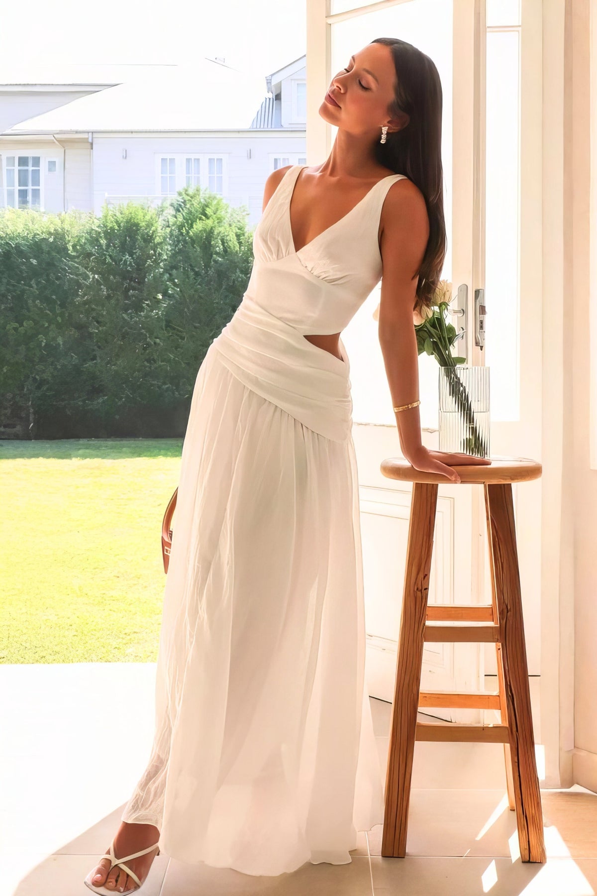 A model leans in an ivory Cutout Back V-Neck Dress with wide straps, ruched waist, side cutout, and a flowing floor-length skirt.