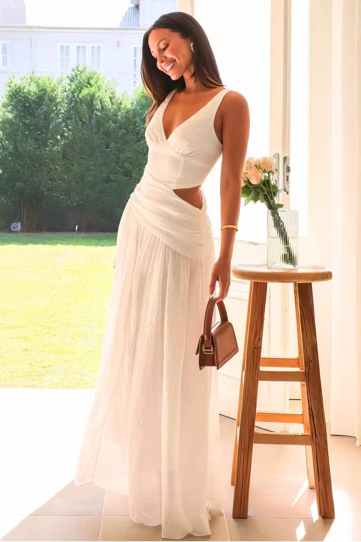 Model leans in an ivory sleeveless V-neck dress with ruched detailing, a waist cutout, fitted bodice, and softly flowing floor-length skirt.