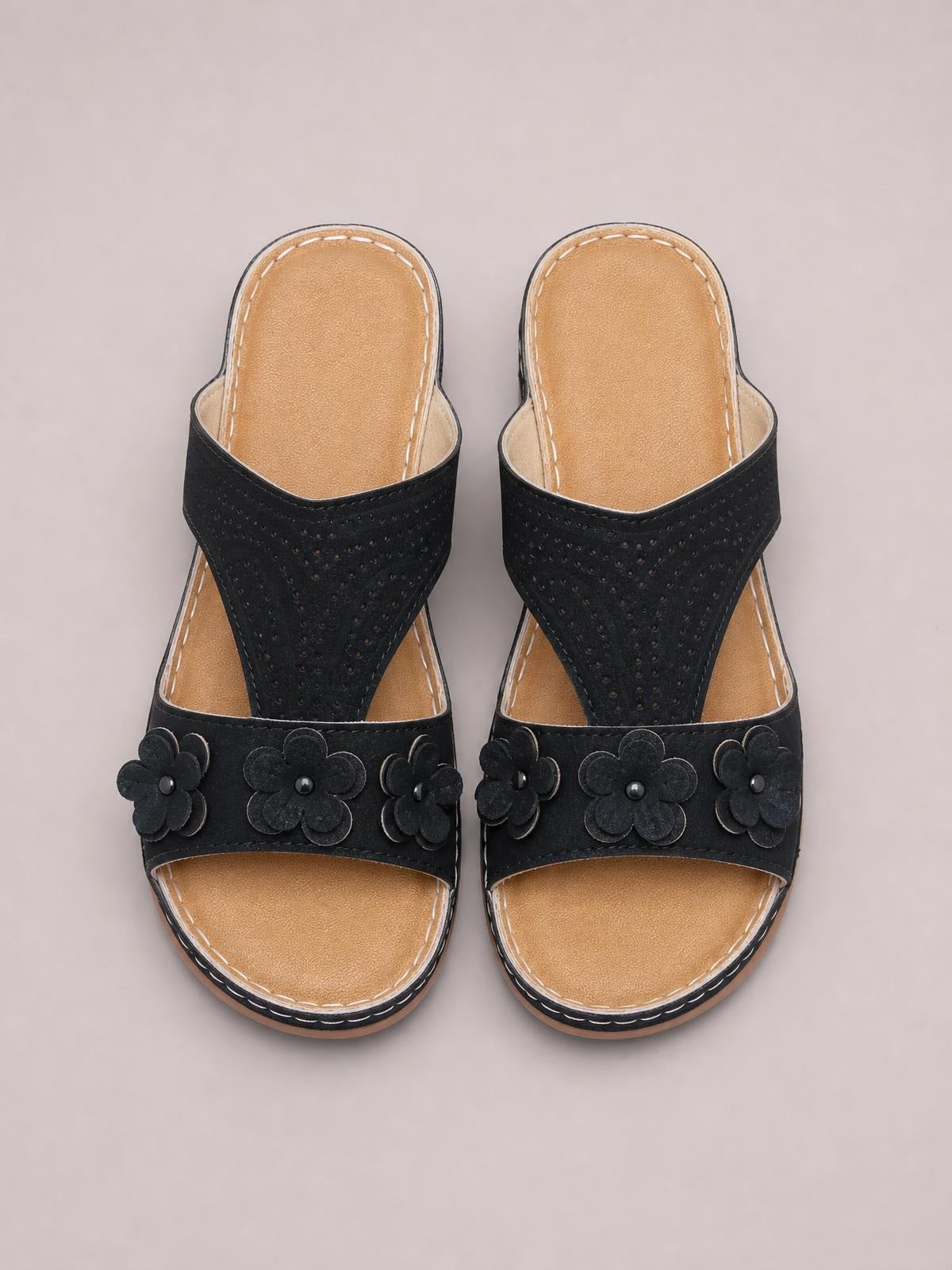 Black cutout flower open-toe flat sandals feature perforated PU leather uppers, floral appliqués, tan footbeds, and stitched rubber soles.