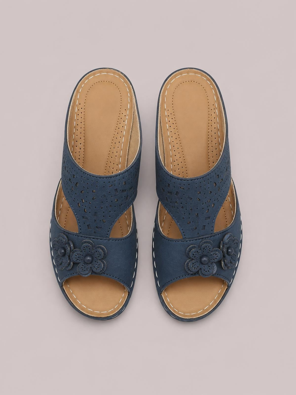 Navy blue open-toe flat sandals feature perforated cutout straps, tan insoles, stitched edges, and layered flower embellishments across the front.