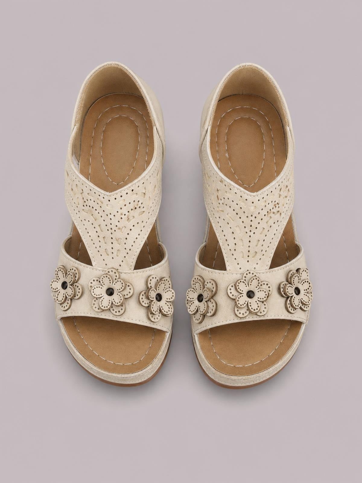Cream open-toe flat sandals feature perforated cutout uppers, wide straps, stitched footbeds, and layered flower embellishments across the toes.