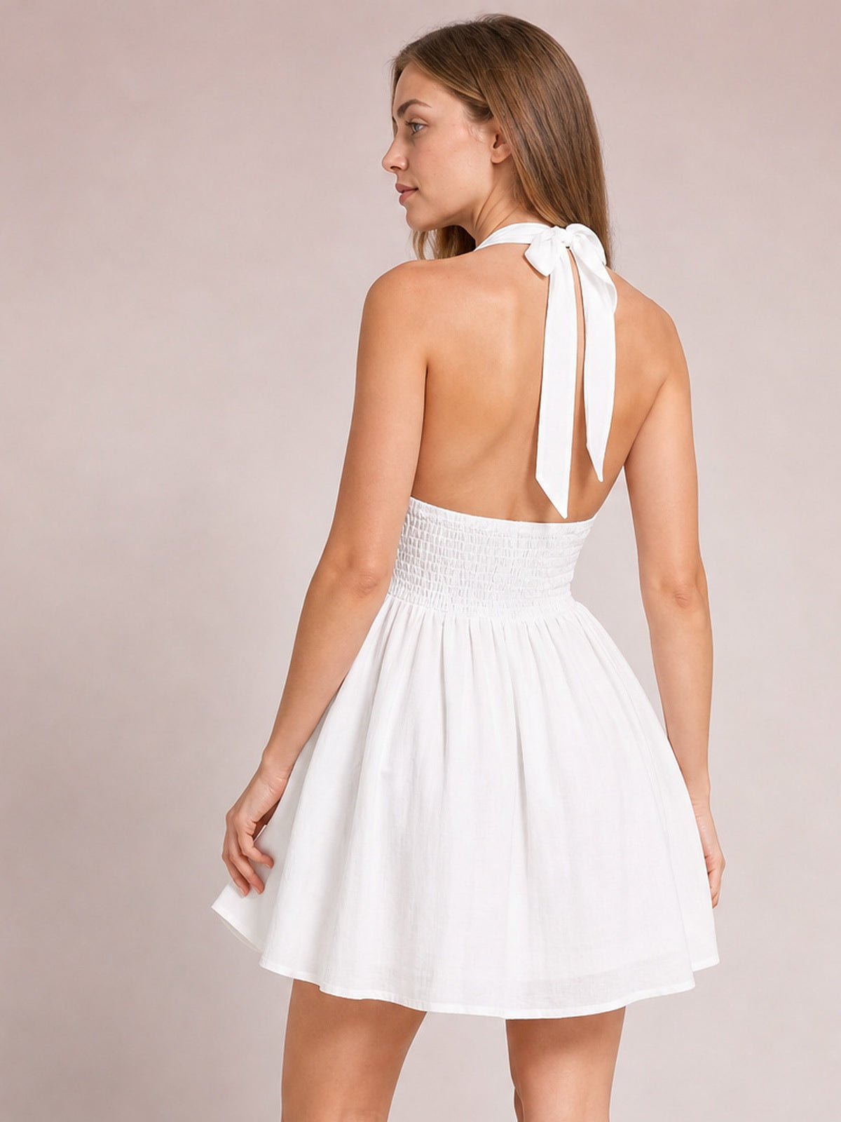 Model poses in a white cutout halter neck mini dress with a tied back, open-back silhouette, ruched waist, and softly flared skirt.