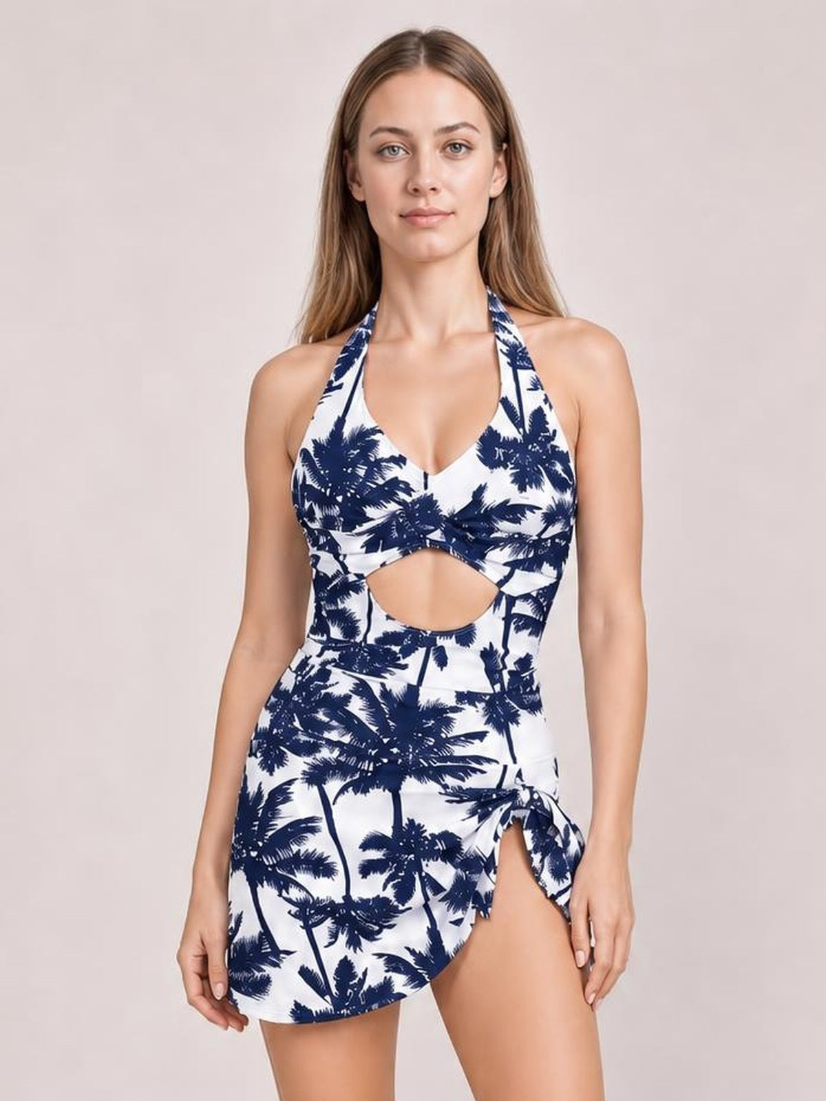 Model standing in a printed white and navy palm-pattern halter neck one-piece swimsuit with a front cutout and asymmetrical side tie detail.