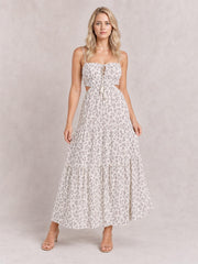 A model stands in a white floral-printed sleeveless cami midi dress with thin straps, a ruched smocked bodice, waist cutouts, front tie, and tiered skirt.