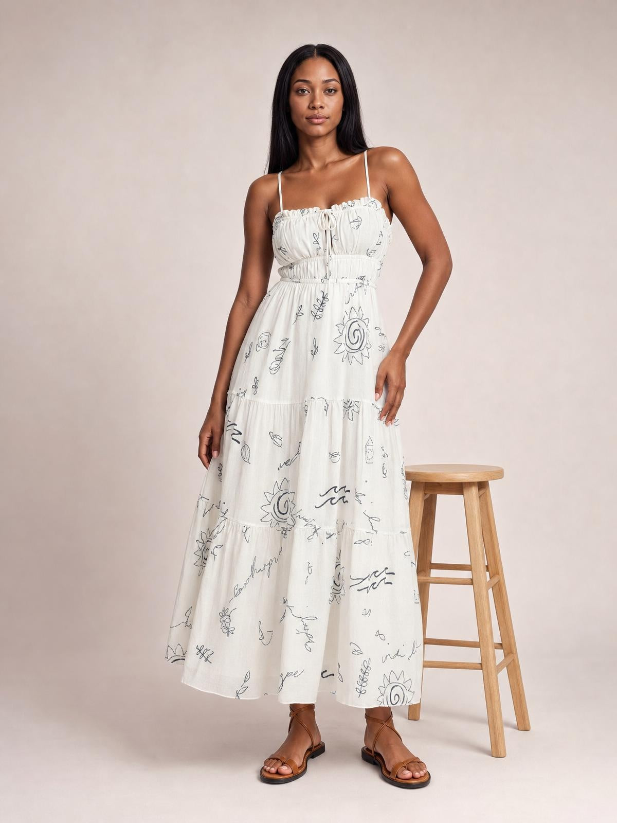 Model wears a white printed sleeveless cami midi dress with delicate straps, ruched smocked bodice, front tie, defined waist, and fluid tiered skirt.