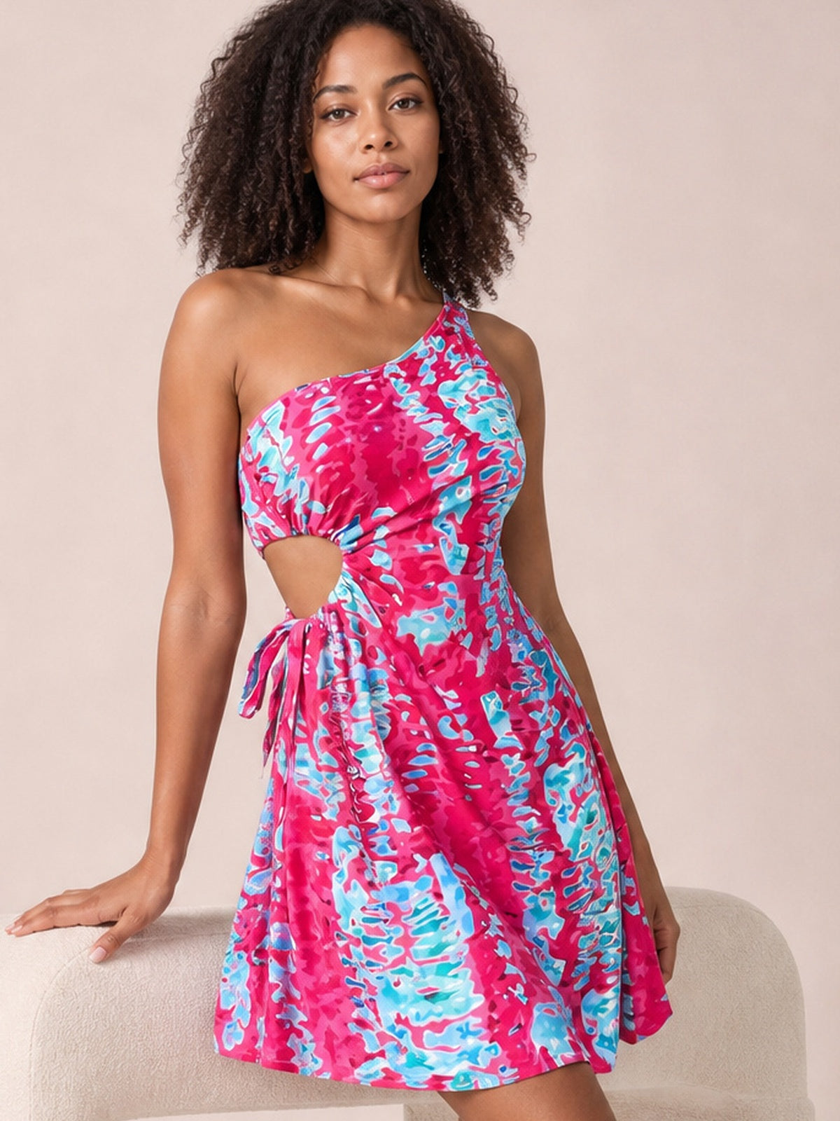Model poses in a vibrant pink and blue printed sleeveless mini dress with an asymmetric one-shoulder neckline, waist cutout, tie detail, and flared skirt.