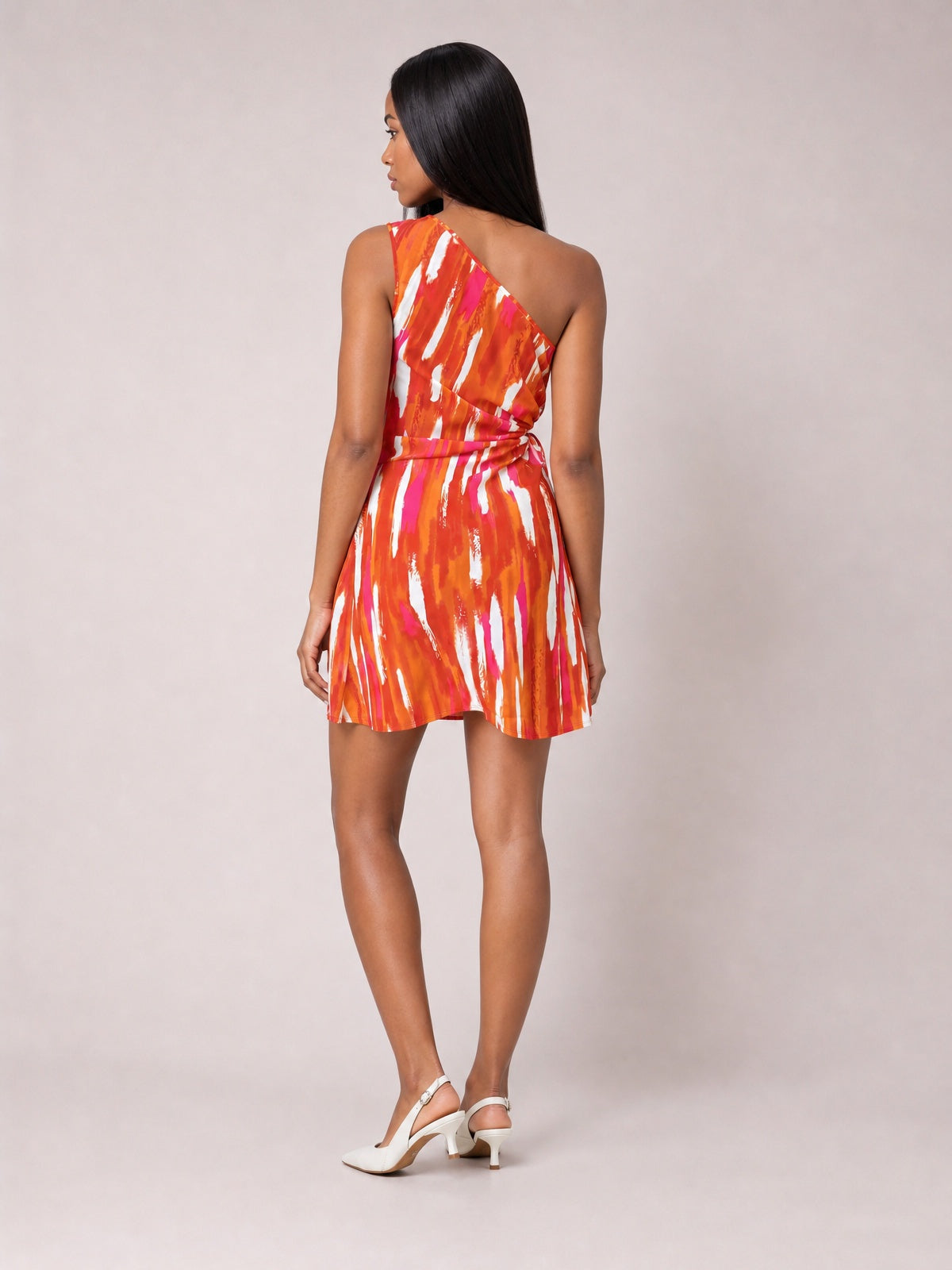 Model shown from behind in a vibrant orange, pink, and white printed sleeveless mini dress with an asymmetric one-shoulder neckline, waist tie, and flared skirt.