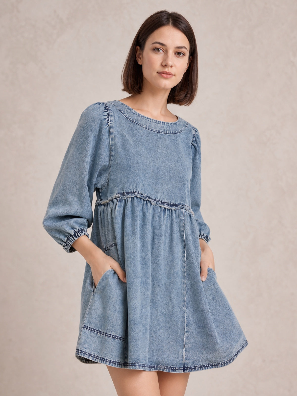 Model poses with hands in the pockets of a light blue denim dress featuring a round neck, gathered waist, balloon sleeves, gathered cuffs, visible seams, and a flared mini silhouette.