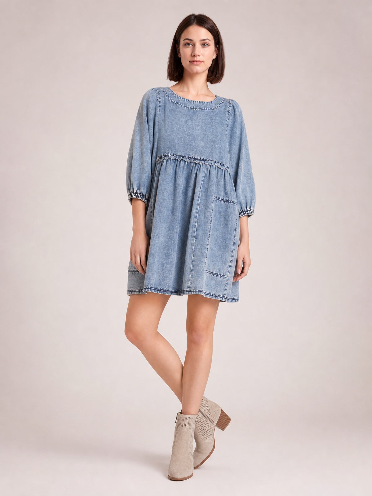 A model poses in a light blue denim mini dress with a round neck, balloon sleeves, gathered waist, flared silhouette, visible seams, and patch pockets.
