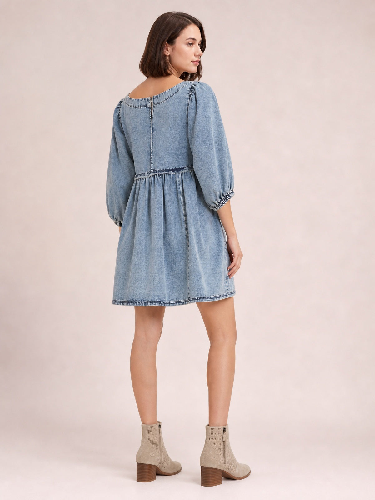 Model shown from the back in a light denim mini dress with a round neck, gathered waist, flared silhouette, balloon sleeves, and cuff details.