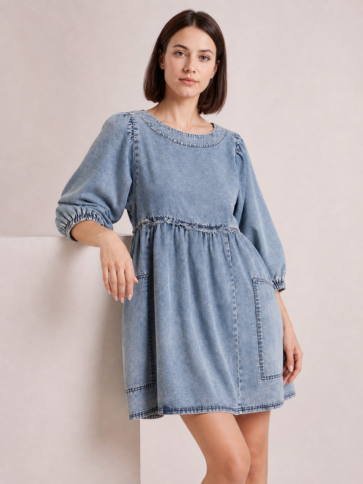 Model wears a light-wash denim dress with a round neck, balloon sleeves, gathered waist, flared mini silhouette, contrast seams, and side pockets.