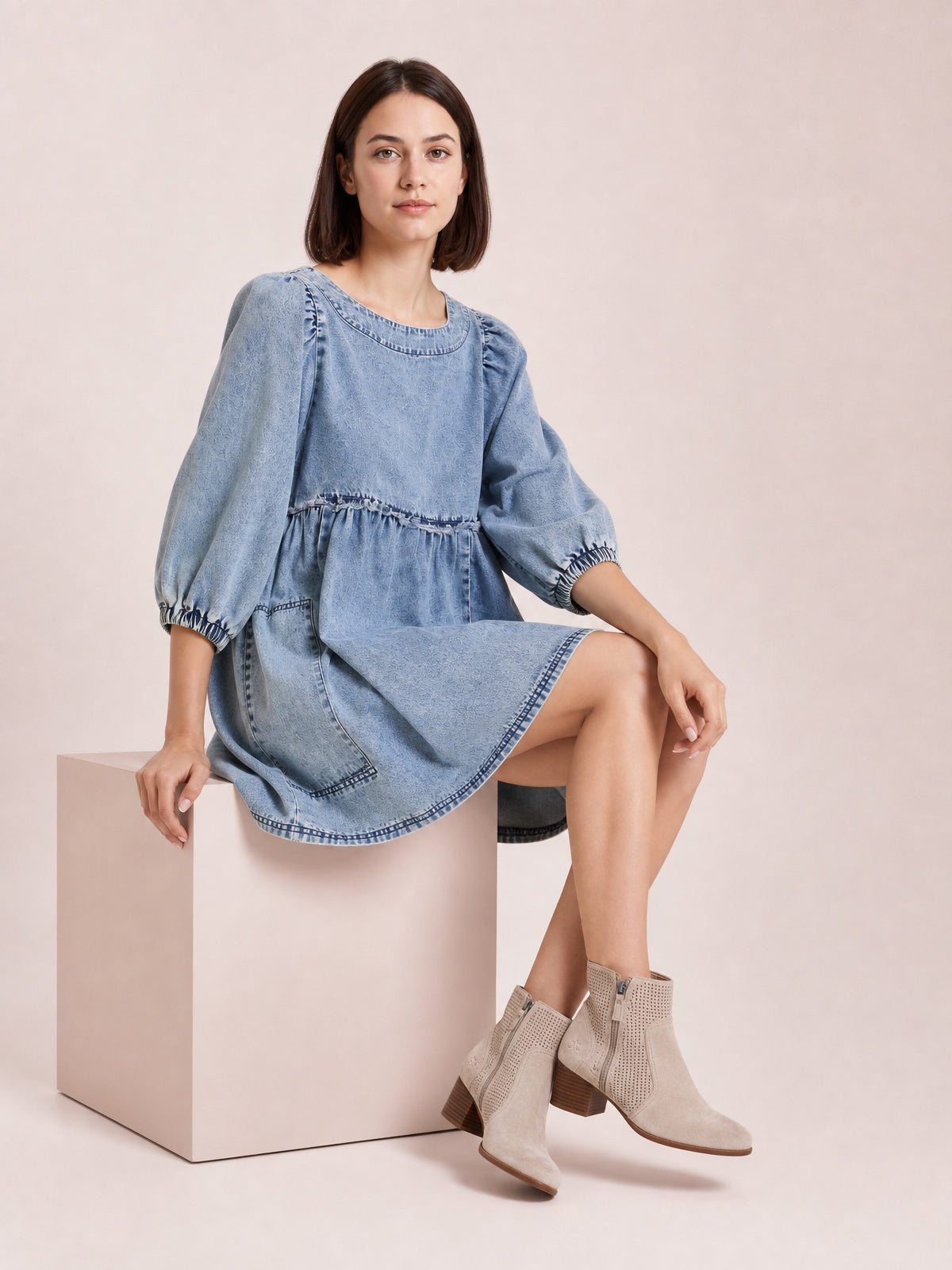 A seated model wears a light denim round-neck dress with voluminous balloon sleeves, gathered cuffs and waist, side pockets, and a gently flared mini silhouette.