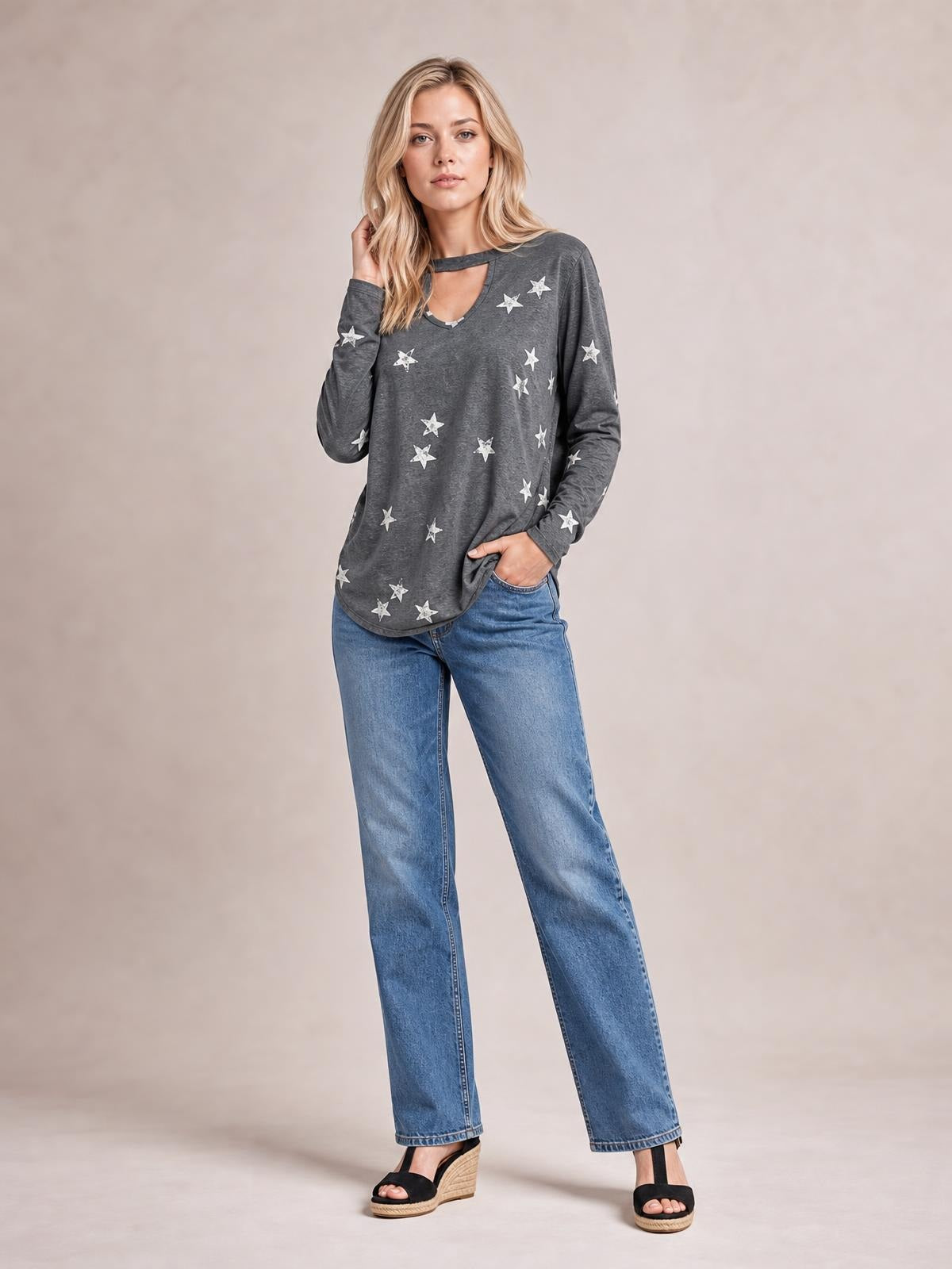 A model wears a charcoal gray cutout round-neck long-sleeve T-shirt with a relaxed silhouette, curved hem, and scattered white stars, posing with one hand in her pocket.