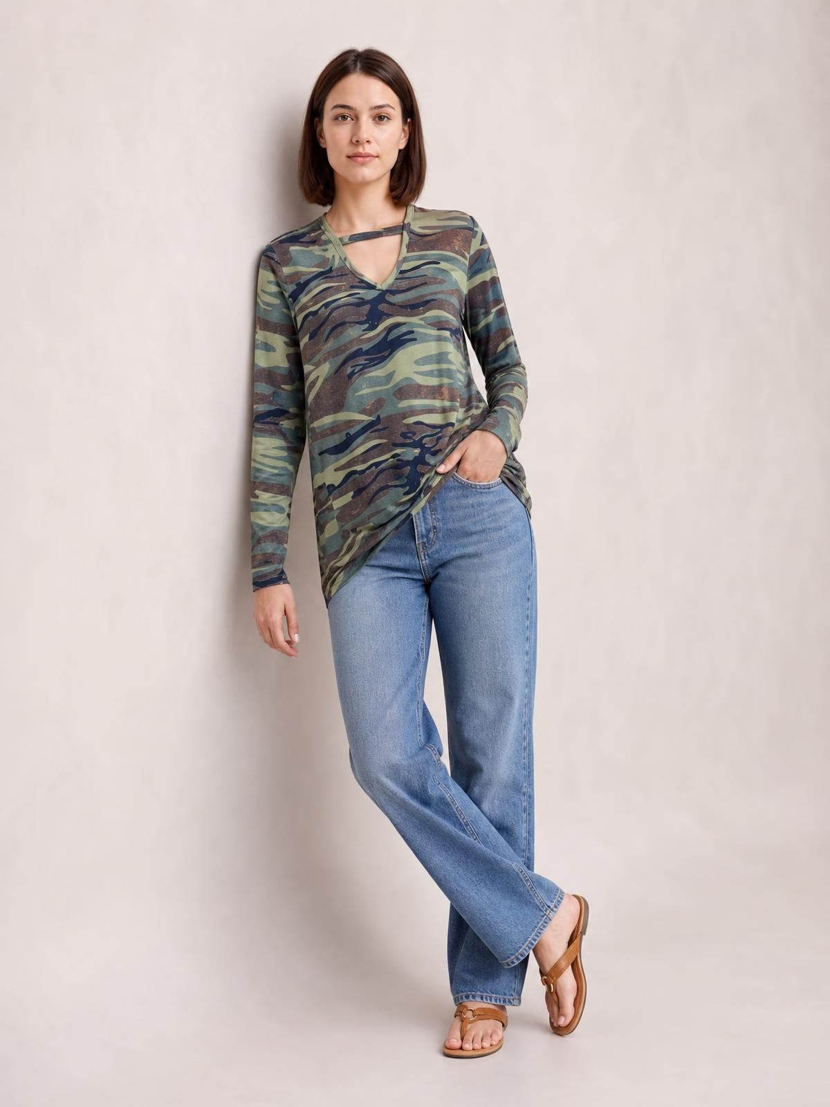 A model poses in a camouflage-print cutout round-neck long-sleeve T-shirt with a straight silhouette, one hand in her pocket and legs crossed.