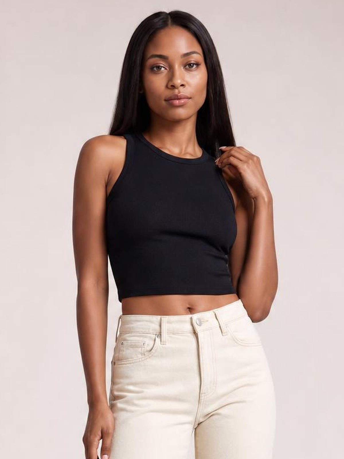 A model wears a fitted black cutout round neck tank with sleeveless construction and a cropped silhouette, posing with one hand near her hair.