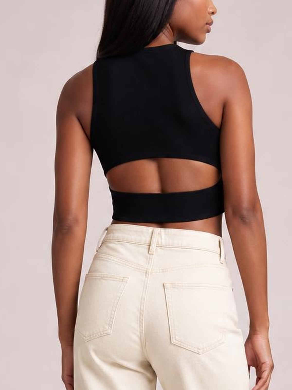 A model wears a black cutout round neck tank with a cropped, fitted silhouette, sleeveless construction, and a wide back opening.
