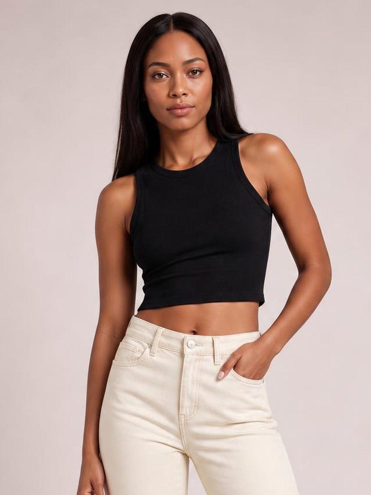 Model wears a black fitted round neck tank with wide shoulder straps and a cropped silhouette, posing with one hand in her jeans pocket.